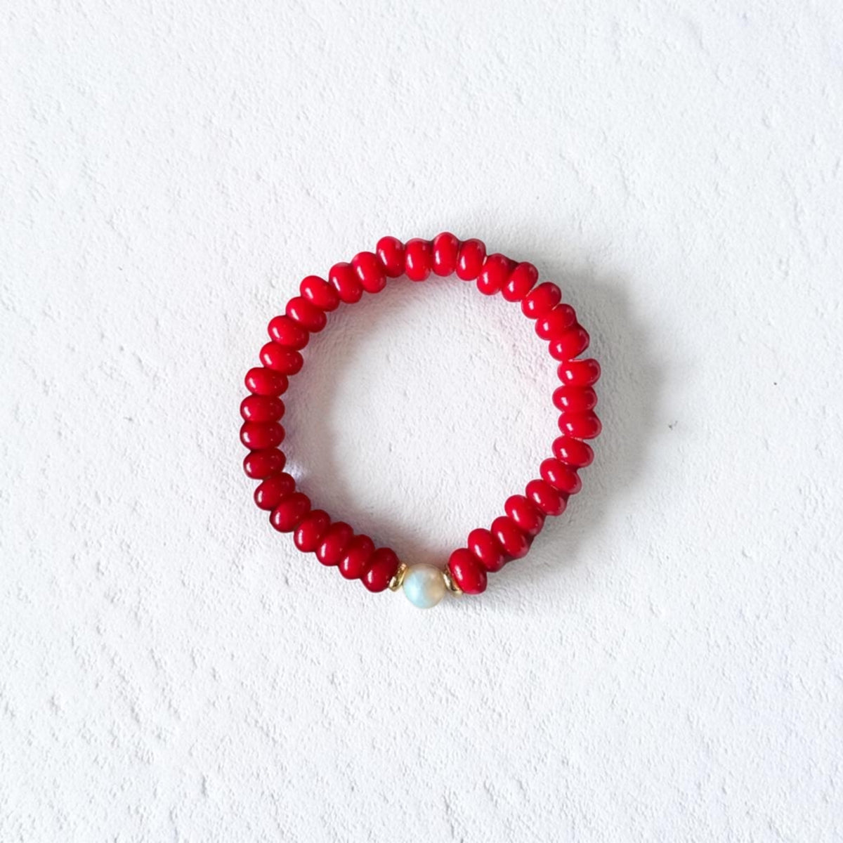 A red beaded bracelet with a small white pearl accent, displayed on a textured white background.