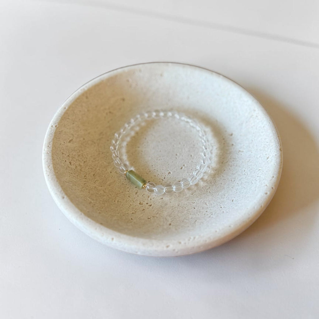 A small round ceramic dish with a speckled off-white glaze, holding a delicate beaded bracelet with a light green bead, set against a plain white background.