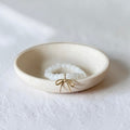 A small round cream-colored ceramic dish holding a white beaded bracelet with a delicate gold bow charm, set against a textured white background.