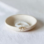 A small round cream-colored ceramic dish holding a white beaded bracelet with a delicate gold bow charm, set against a textured white background.