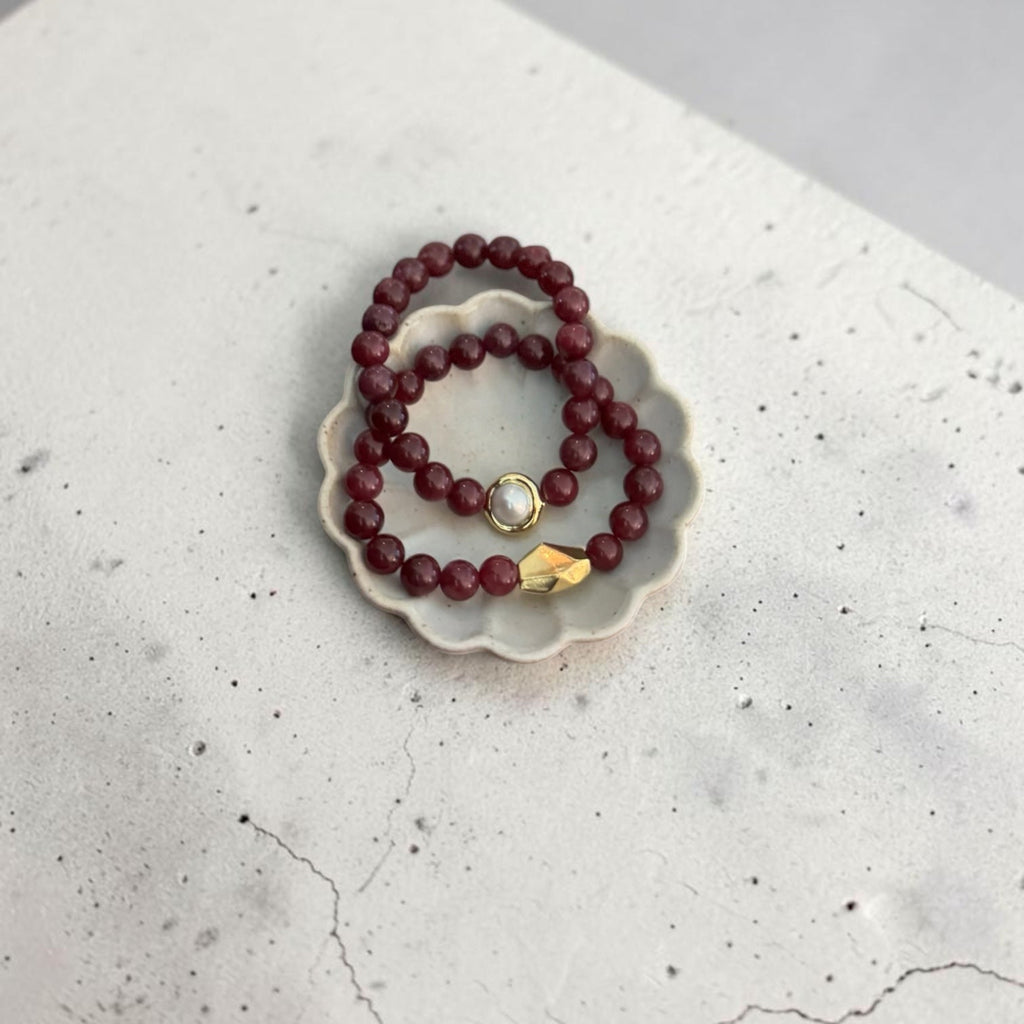 Two beaded bracelets, one dark red and one maroon with a gold-toned bead accent, displayed on a small white ceramic dish over a textured gray surface.