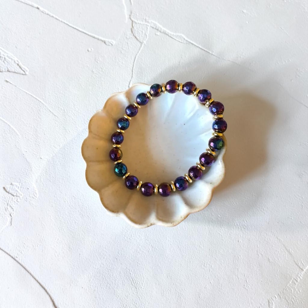 A beaded stretch bracelet with iridescent purple and blue beads separated by gold spacer rings, displayed on a small white scalloped dish against a textured white background.