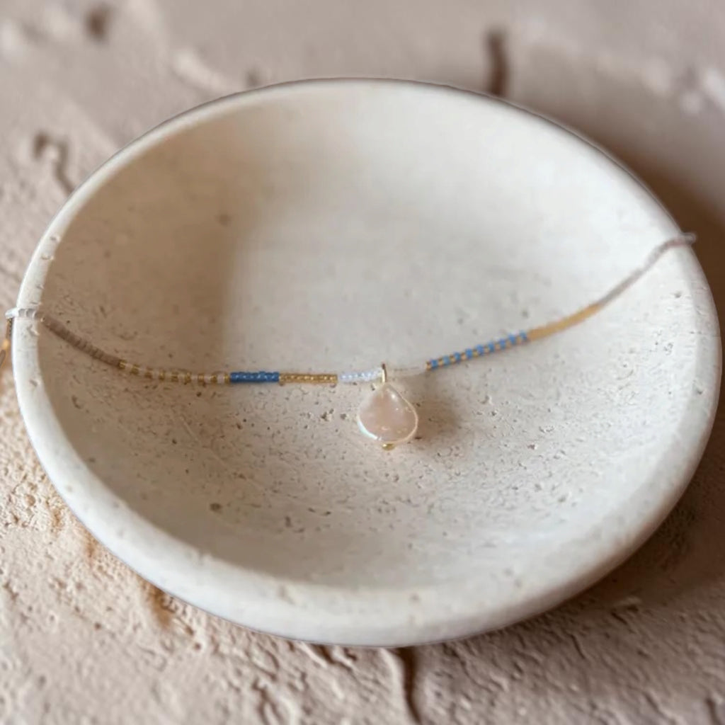 A delicate beaded necklace with a teardrop-shaped pale pink stone pendant, displayed on a textured beige ceramic dish.