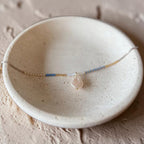 A delicate beaded necklace with a teardrop-shaped pale pink stone pendant, displayed on a textured beige ceramic dish.