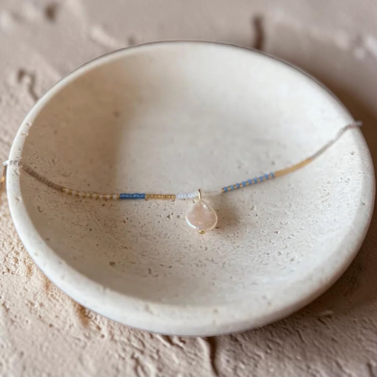 A delicate beaded necklace with a teardrop-shaped pale pink stone pendant, displayed on a textured beige ceramic dish.