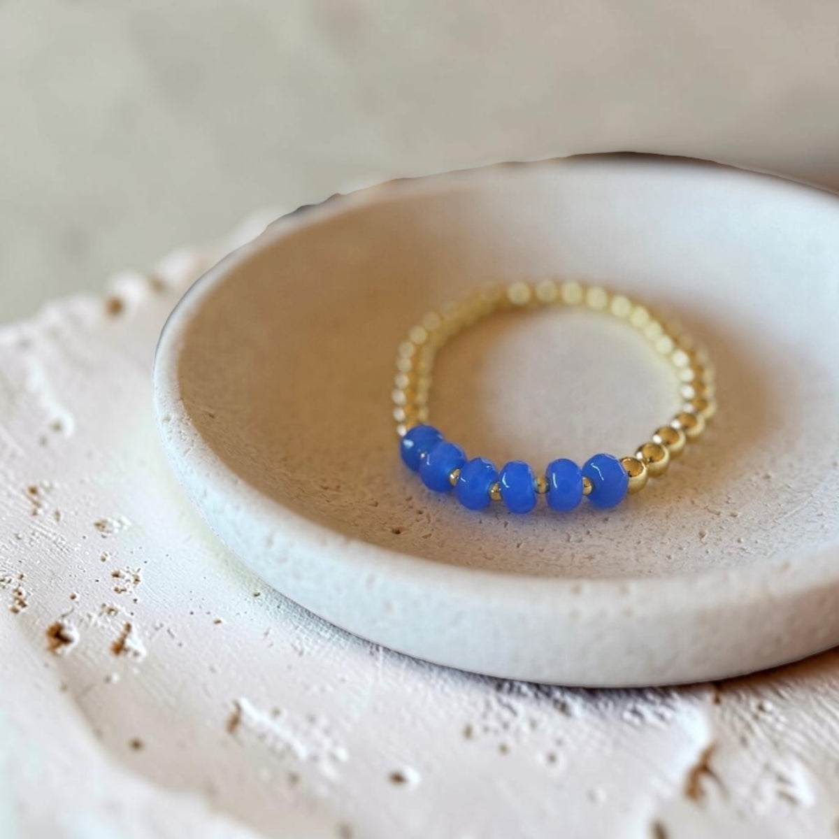 A beaded stretch bracelet featuring five glossy cobalt blue beads separated by small gold spacer beads, displayed on a textured off-white ceramic dish.