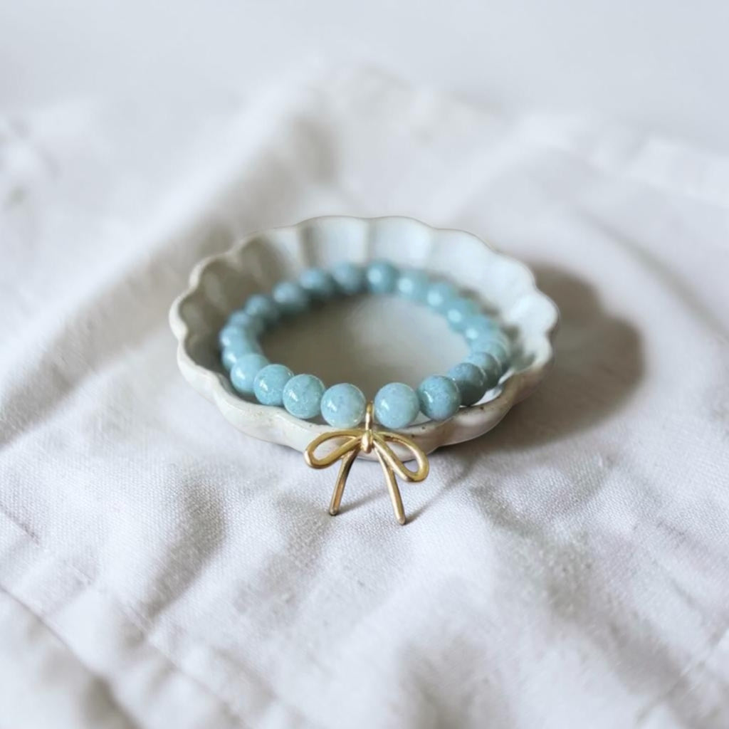 A light blue beaded bracelet with a gold bow charm, displayed inside a small white scalloped dish on a textured white fabric background.