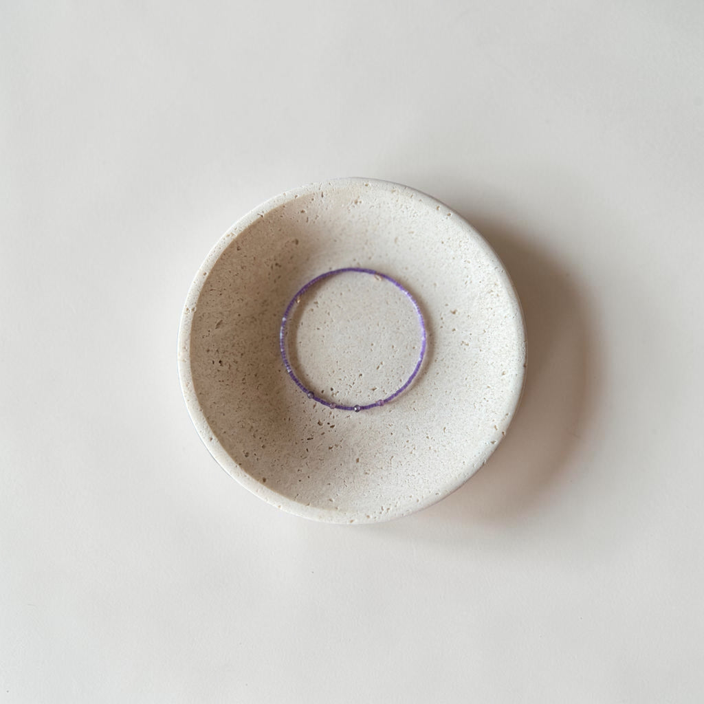 A small round ceramic dish with a speckled off-white glaze and a contrasting purple wire bracelet, photographed from directly above on a light neutral background.