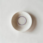 A small round ceramic dish with a speckled off-white glaze and a contrasting purple wire bracelet, photographed from directly above on a light neutral background.