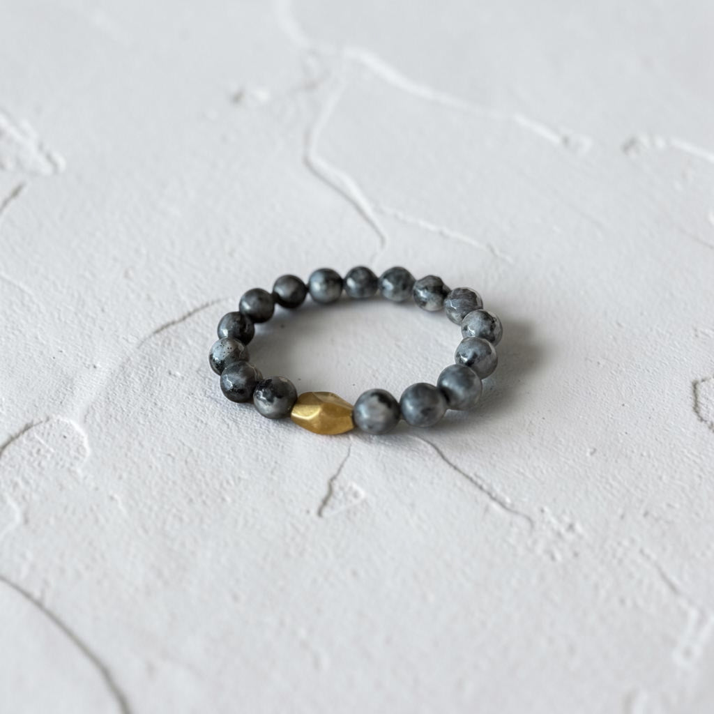 A beaded stretch bracelet featuring dark gray marbled beads and a single faceted gold accent bead, resting on a textured white surface.