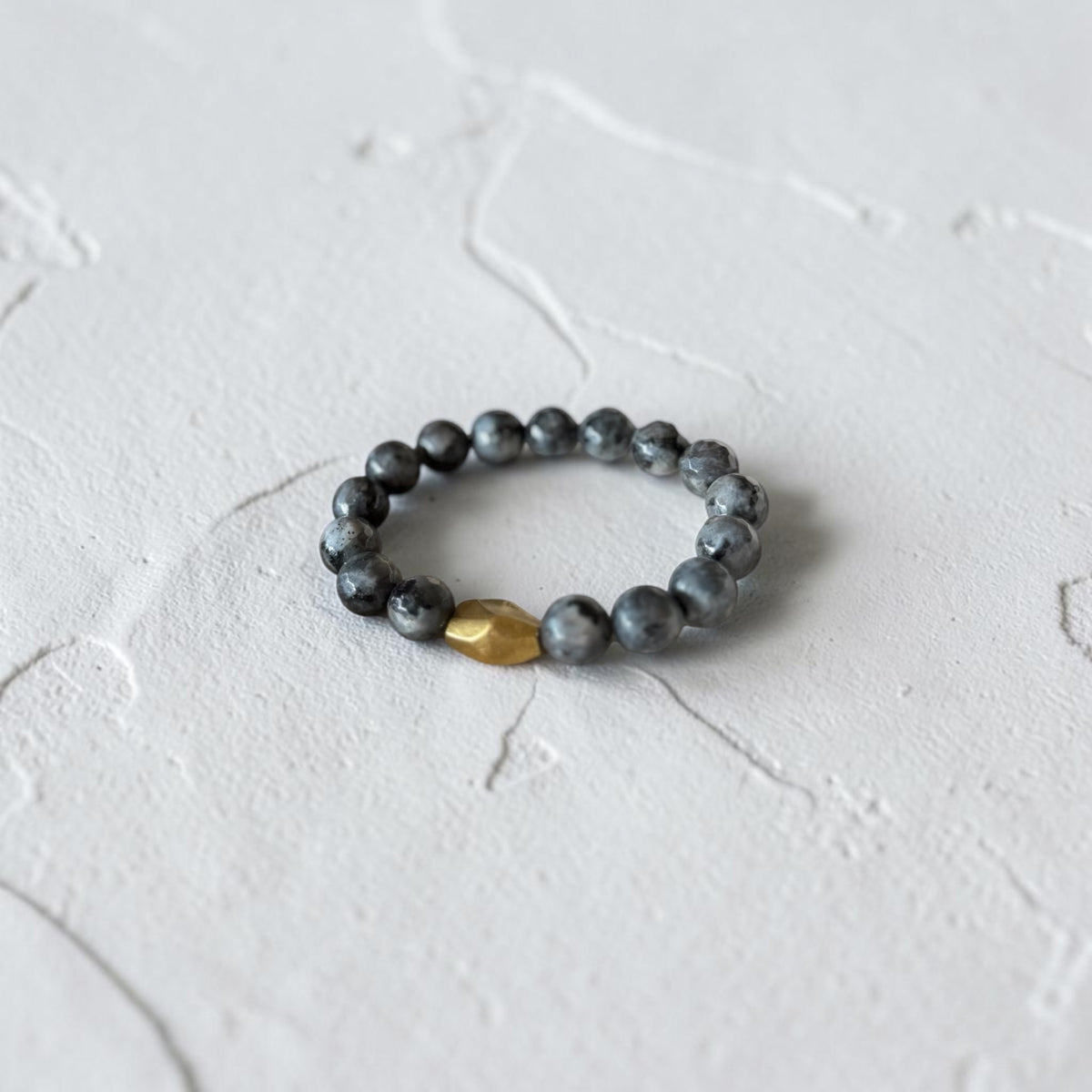 A beaded stretch bracelet featuring dark gray marbled beads and a single faceted gold accent bead, resting on a textured white surface.