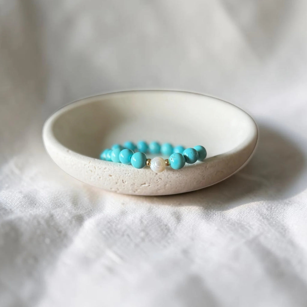 A beaded bracelet with turquoise-blue beads and a small white bead separated by gold foil, resting inside a small round off-white ceramic dish on a white fabric background.