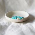 A beaded bracelet with turquoise-blue beads and a small white bead separated by gold foil, resting inside a small round off-white ceramic dish on a white fabric background.