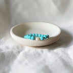 A beaded bracelet with turquoise-blue beads and a small white bead separated by gold foil, resting inside a small round off-white ceramic dish on a white fabric background.