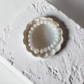 A white stripe beaded bracelet with a single pearl-like bead, displayed inside a scalloped cream ceramic dish on a textured white surface.
