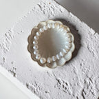 A white stripe beaded bracelet with a single pearl-like bead, displayed inside a scalloped cream ceramic dish on a textured white surface.