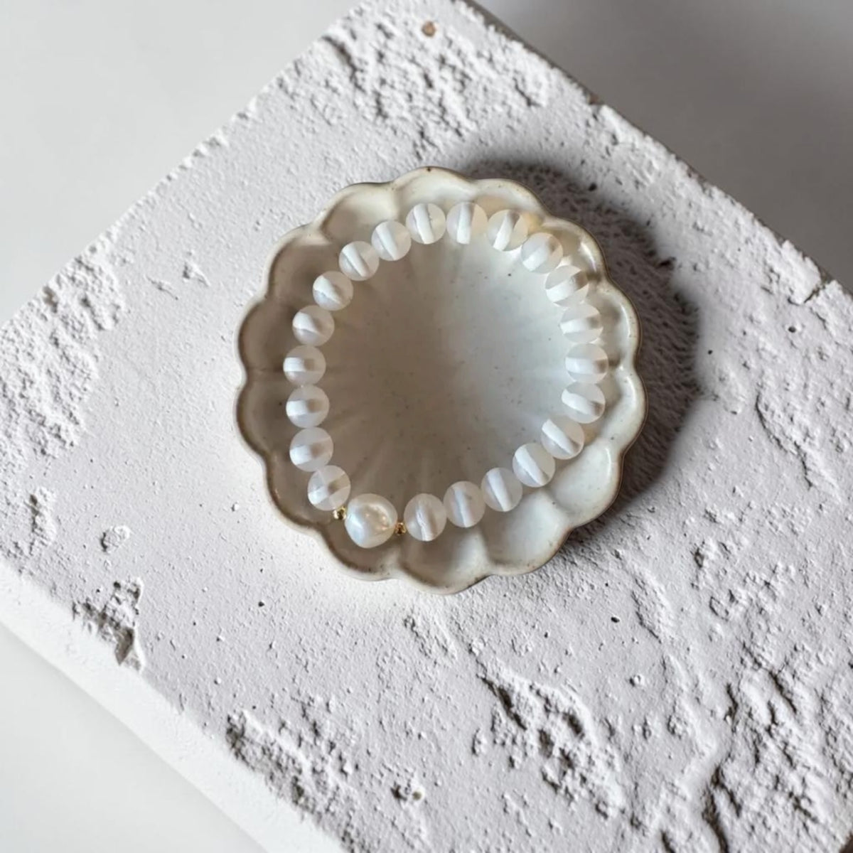 A white stripe beaded bracelet with a single pearl-like bead, displayed inside a scalloped cream ceramic dish on a textured white surface.