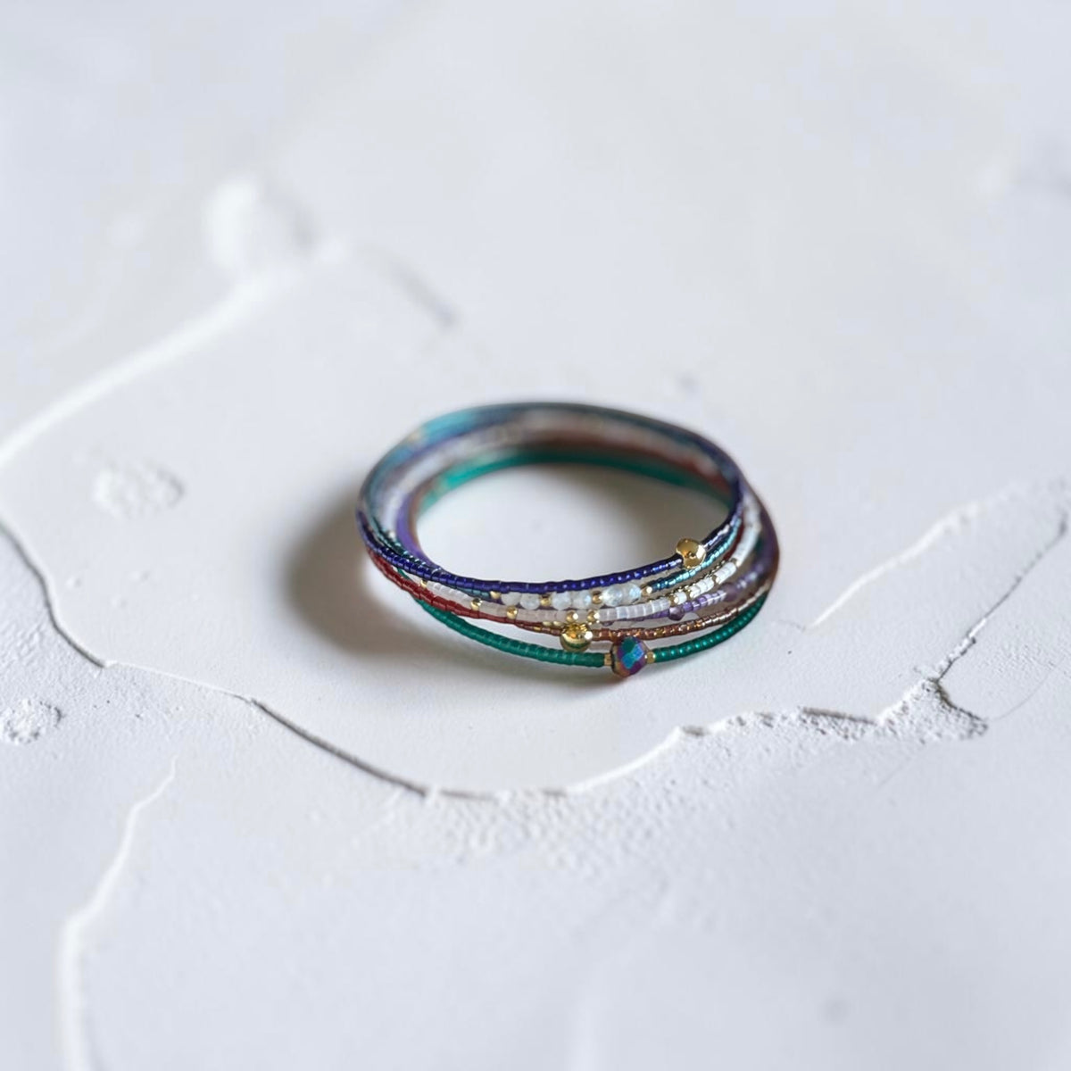 A delicate multi-layered bracelets with colorful glass strands, tiny white pearls, and small gold beads, resting on a textured white surface.