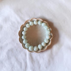 A beaded bracelet made of light green oval beads, displayed on a small scalloped ceramic dish over a white fabric background.