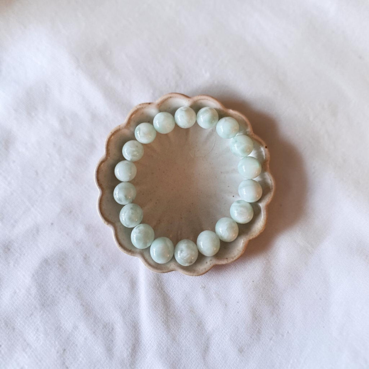 A beaded bracelet made of light green oval beads, displayed on a small scalloped ceramic dish over a white fabric background.