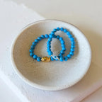 Two beaded stretch bracelets in bright turquoise blue with a small gold and white evil eye charm, displayed in a small speckled ceramic dish.