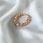 A peach-colored beaded bracelet with a single white pearl and small gold spacer beads, resting on a draped white satin fabric.