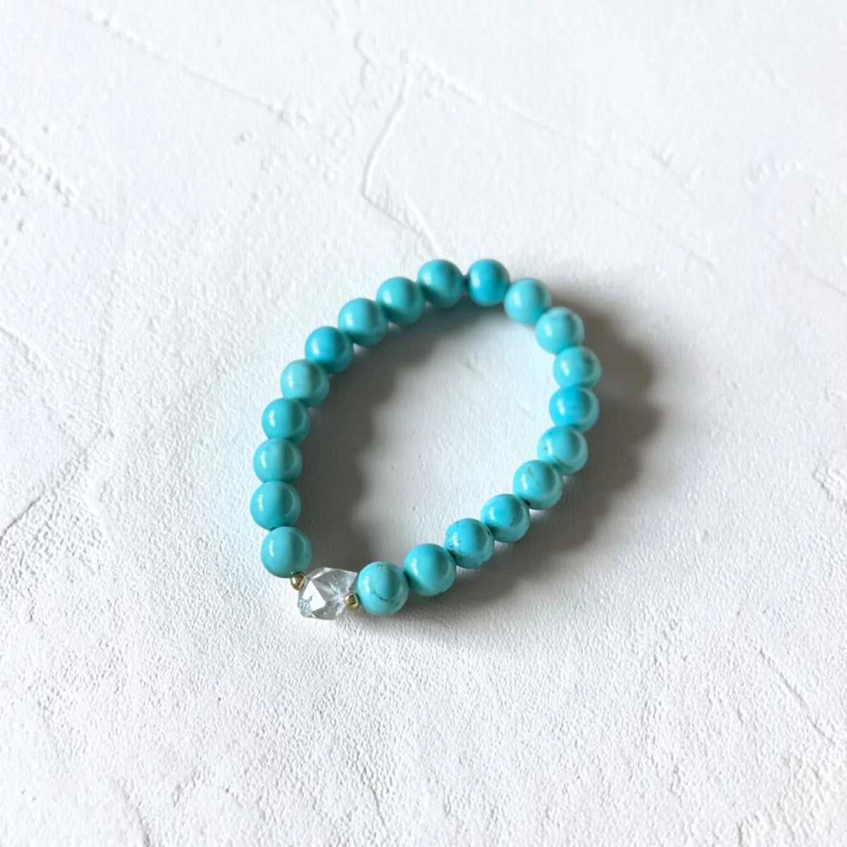 A turquoise beaded stretch bracelet with a small clear faceted accent bead, displayed on a textured white surface.