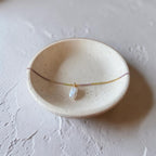 A delicate necklace with a small white irregular stone pendant and a thin gold chain, displayed on a speckled beige ceramic dish against a textured light gray background.