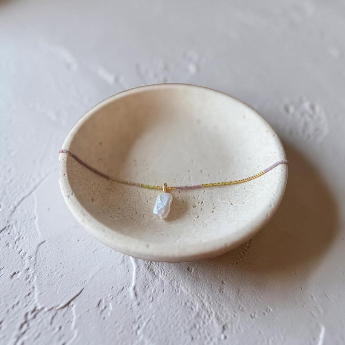 A delicate necklace with a small white irregular stone pendant and a thin gold chain, displayed on a speckled beige ceramic dish against a textured light gray background.
