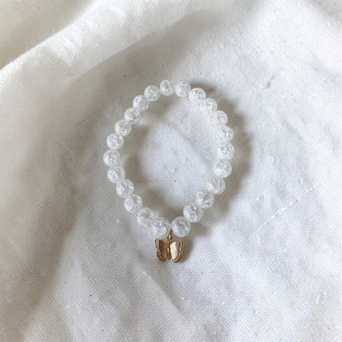 A beaded stretch bracelet made of clear, faceted round beads with a small gold butterfly charm, resting on a textured white fabric background.