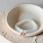A beaded bracelet with translucent white round beads and a small gold butterfly charm, displayed inside a shallow cream ceramic dish.