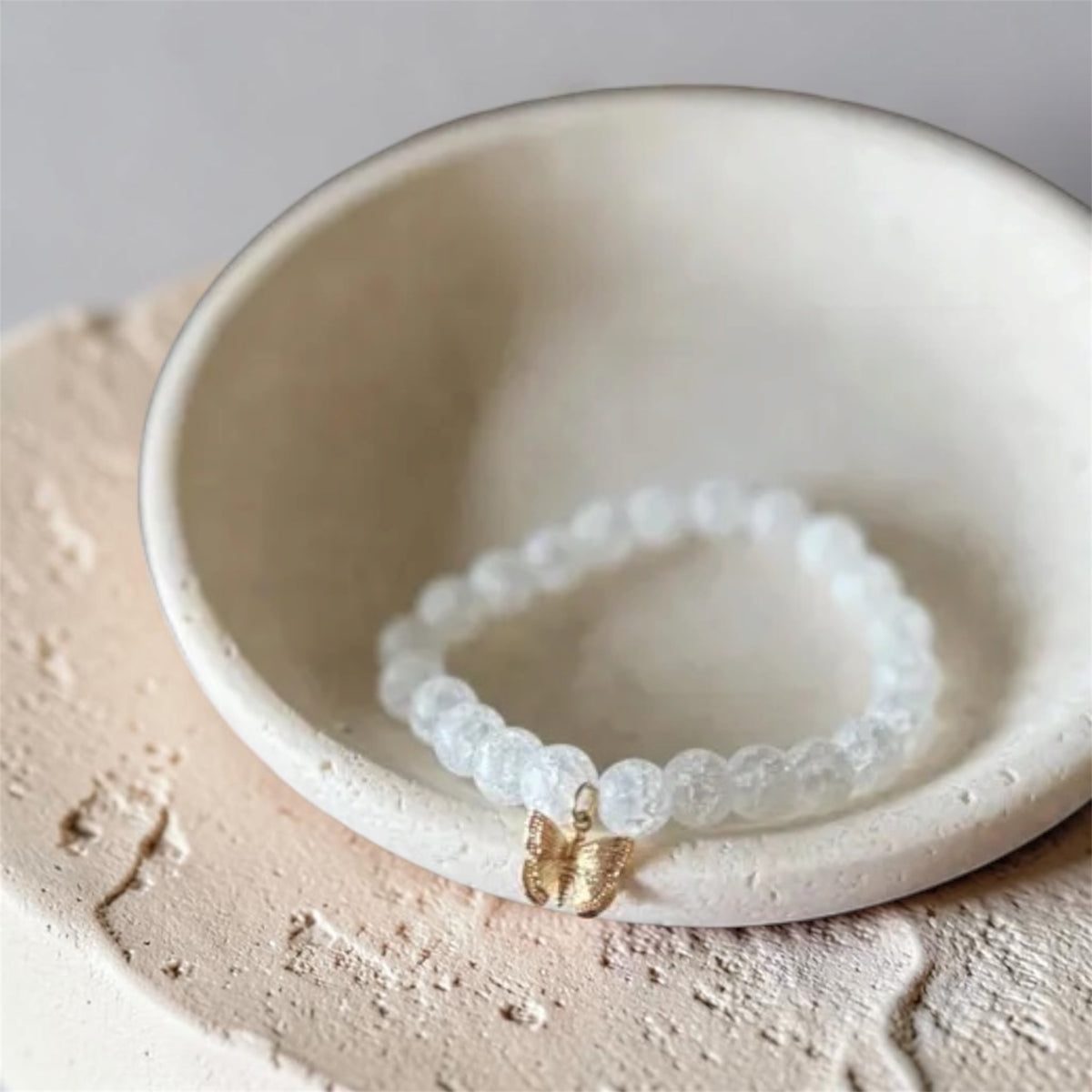 A beaded bracelet with translucent white round beads and a small gold butterfly charm, displayed inside a shallow cream ceramic dish.