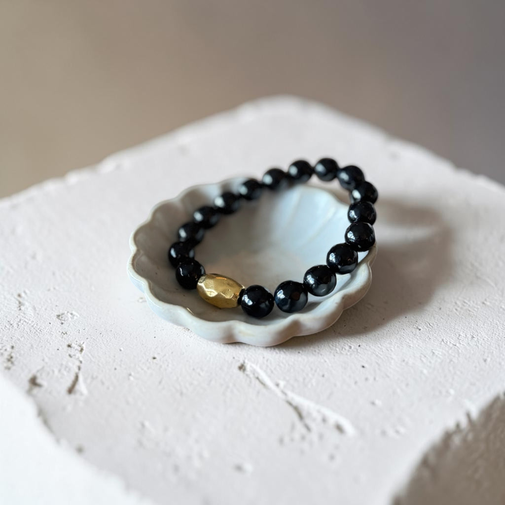 A beaded bracelet with glossy black round beads and a single textured gold accent bead, displayed on a small white ceramic dish atop a rough white stone block.