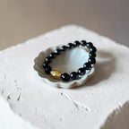 A beaded bracelet with glossy black round beads and a single textured gold accent bead, displayed on a small white ceramic dish atop a rough white stone block.