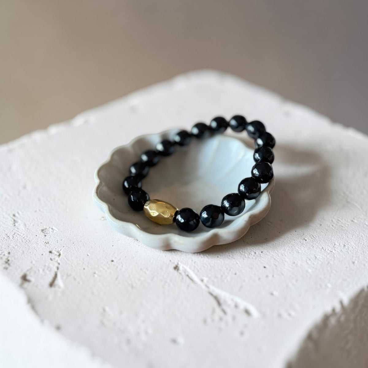 A beaded bracelet with glossy black round beads and a single textured gold accent bead, displayed on a small white ceramic dish atop a rough white stone block.