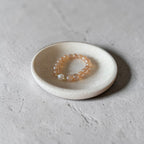 A beaded bracelet with peach-orange translucent beads and a small white pearl accent, displayed on a small round ceramic dish over a textured gray concrete surface.