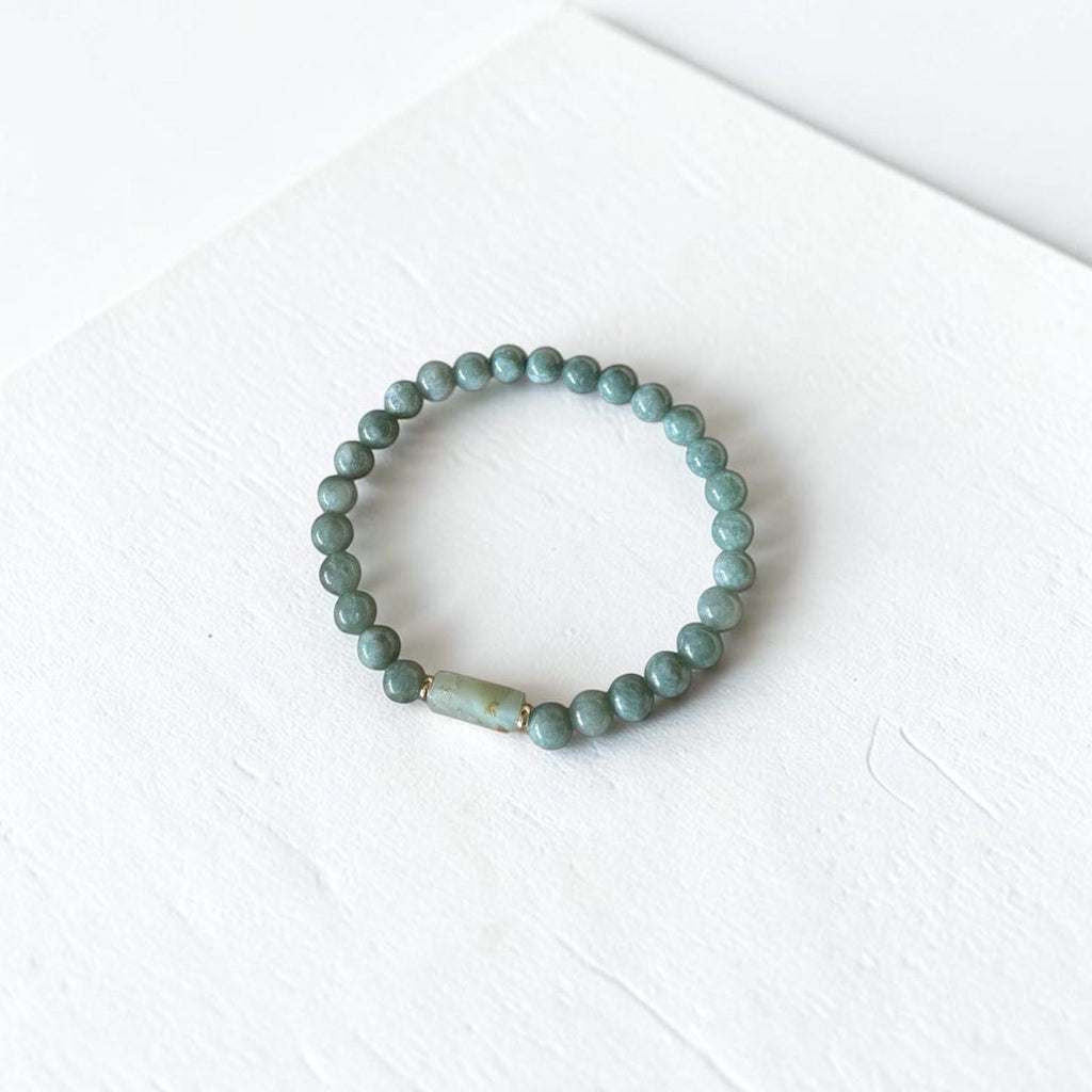 A beaded stretch bracelet made of small round sage green beads with a single larger, textured light green accent bead, displayed on a white textured surface.