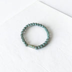 A beaded stretch bracelet made of small round sage green beads with a single larger, textured light green accent bead, displayed on a white textured surface.