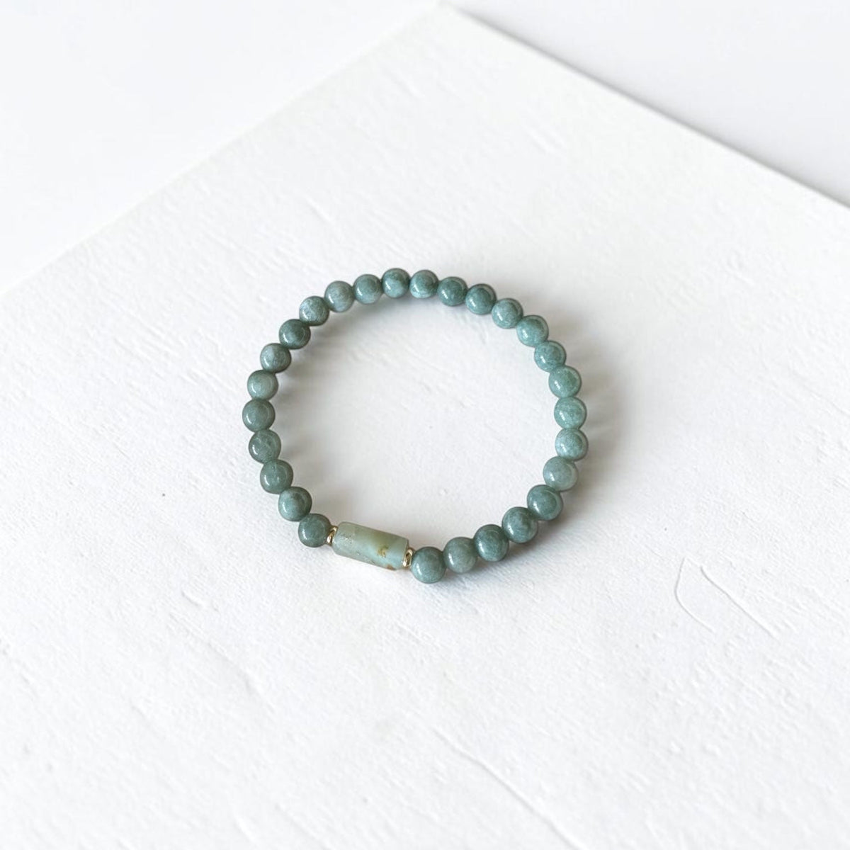 A beaded stretch bracelet made of small round sage green beads with a single larger, textured light green accent bead, displayed on a white textured surface.
