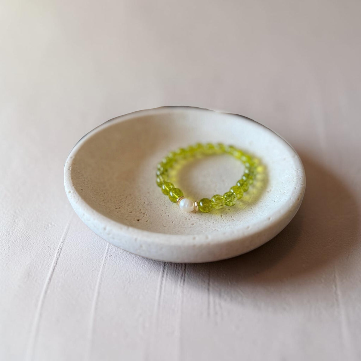 A beaded stretch bracelet in translucent light green with a small white pearl accent, displayed inside a small speckled ceramic dish on a light beige textured surface.
