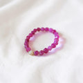 A beaded stretch bracelet made of faceted magenta-pink beads with a small white pearl accent, resting on a soft white fabric background.