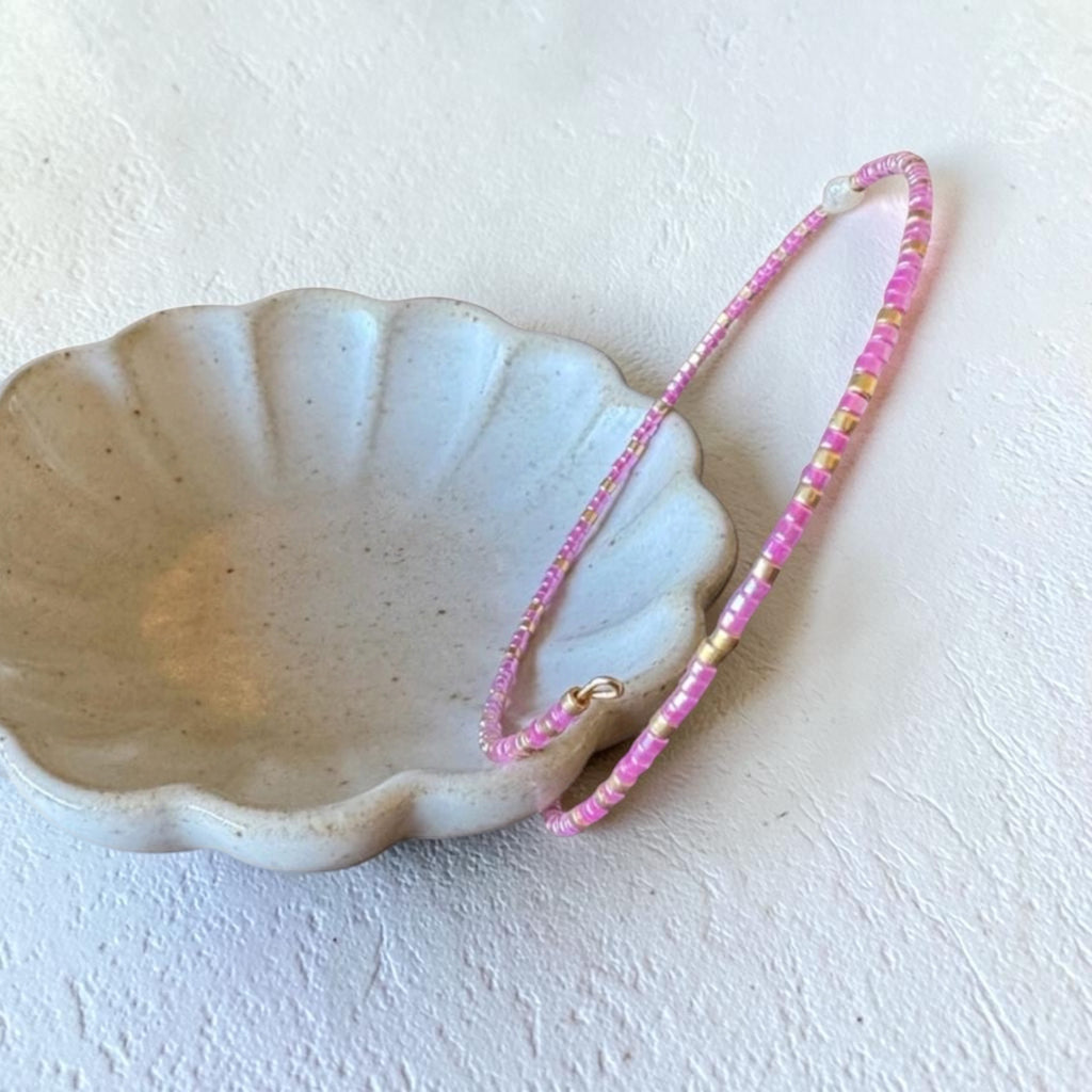 A delicate pink beaded bracelet with small gold spacer beads and a gold clasp, draped over a small scalloped ceramic dish on a textured off-white surface.
