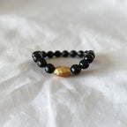 A beaded stretch bracelet featuring glossy black round beads and a single hammered gold oval accent bead, displayed on a textured white fabric background.