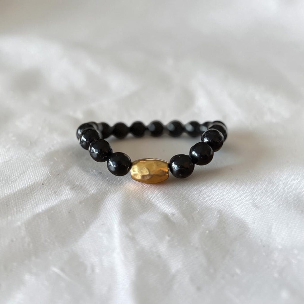 A beaded stretch bracelet featuring glossy black round beads and a single hammered gold oval accent bead, displayed on a textured white fabric background.