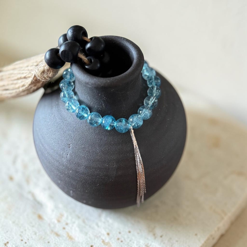 A beaded bracelet with light blue faceted beads and a silver-toned tassel, draped around a matte black ceramic vase. The necklace also features a cluster of matte black beads at the top, resting on a light beige textured surface.