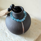 A beaded bracelet with light blue faceted beads and a silver-toned tassel, draped around a matte black ceramic vase. The necklace also features a cluster of matte black beads at the top, resting on a light beige textured surface.