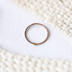 A minimalist bracelet made of textured copper-colored metal with a small gold bead accent, resting on a white surface with a soft-focus woven cream background.