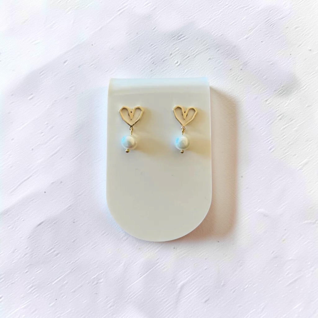 A pair of gold heart-shaped drop earrings with small white pearl-like beads, displayed on a translucent white stand against a textured white background.