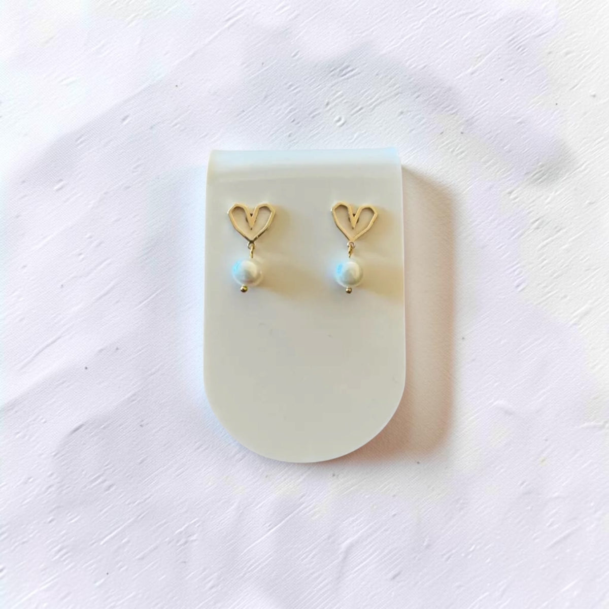A pair of gold heart-shaped drop earrings with small white pearl-like beads, displayed on a translucent white stand against a textured white background.