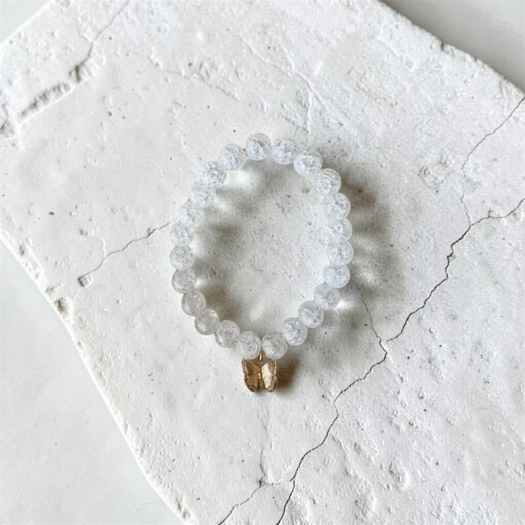 A beaded stretch bracelet made of clear, faceted round beads with a small gold-toned dangling charm, displayed on a textured white stone slab.